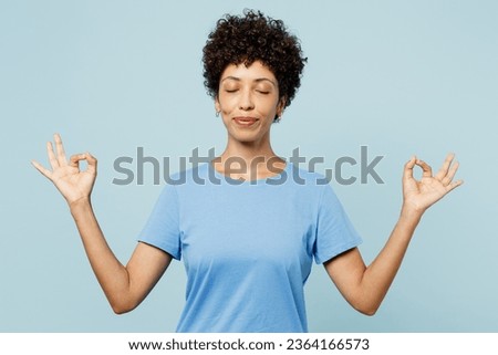 Young spiritual woman of African American ethnicity wear t-shirt casual clothes hold spreading hands in yoga om aum gesture relax meditate try to calm down isolated on plain light blue cyan background