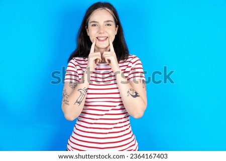 Happy Young beautiful woman wearing striped T-Shirt over blue studio background with toothy smile, keeps index fingers near mouth, fingers pointing and forcing cheerful smile