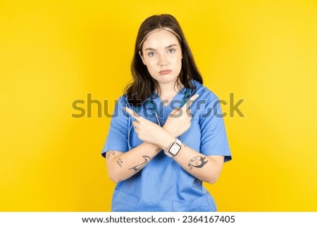 Serious young beautiful doctor woman standing over yellow background crosses hands and points at different sides hesitates between two items. Hard decision concept