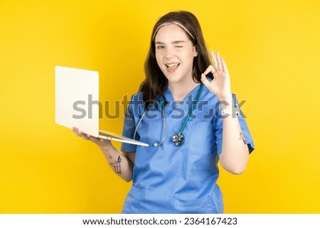 Attractive cheerful skilled young beautiful doctor woman standing over blue  background using laptop showing ok-sign winkin