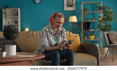 A woman wearing red wireless headphones and holding a phone is sitting on the couch at home.