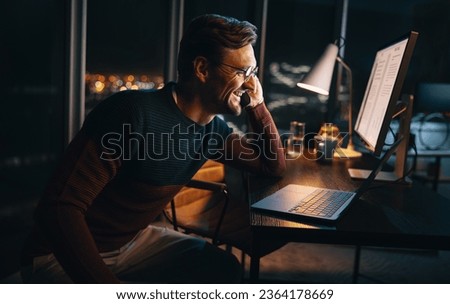 A happy business person works late at night in the office, discussing a project on a phone call. He sits at his desk, focused on his laptop, maintaining a professional work-life balance.