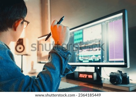 Asian male creator wearing a denim jacket, works with footage or video on his personal computer, in his creative office studio.