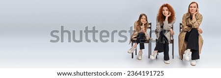 female generations, happy redhead women and child sitting in row on chairs on grey backdrop Royalty-Free Stock Photo #2364191725