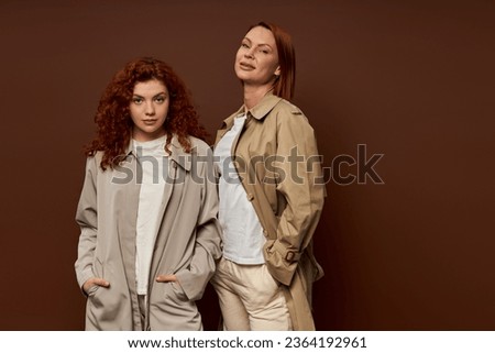 two generations, redhead women in trendy autumn attire posing on brown background, trench coats