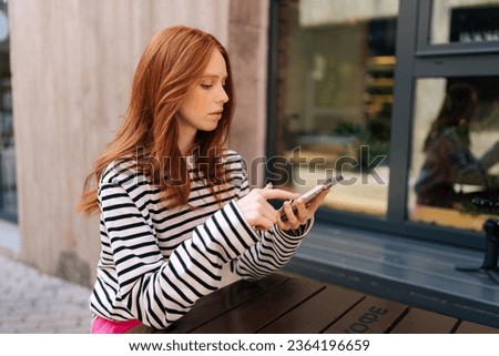 Side view of pensive young woman looking at phone online dating swiping left and right on a dating app, liking some of profiles and not liking some, sitting at table in outdoor cafe. Royalty-Free Stock Photo #2364196659