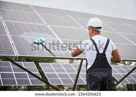 Worker cleaning solar photovoltaic panel. Man technician making sure solar batteries in good condition. Back view of professional worker in overalls and helmet cleaning panel by mop. Royalty-Free Stock Photo #2364206623