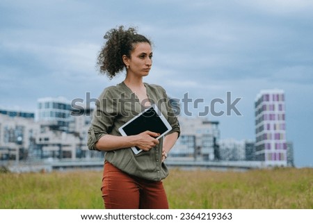 Purposeful curly female realtor in casual holding digital tablet stands on meadow against city buildings , thinks about new construction project, Focused woman, architect planning new block.