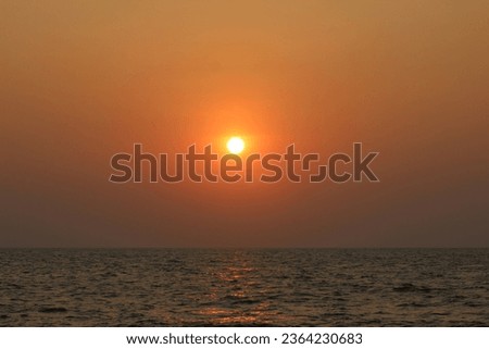 Beautiful photo of a sunset over the ocean. The sun is a large orange ball in the sky, casting a warm glow over the horizon. The ocean is a deep blue with small waves. The sky is a gradient of orange. Royalty-Free Stock Photo #2364230683