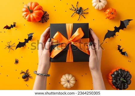 Elevate Halloween gifting for close ones. Above view features a woman's hands with a black present box with orange bow and Halloween decor on an orange isolated backdrop, ready for adverts or text