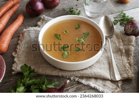 Chicken bone broth in a white soup bowl on a wooden table with fresh vegetables and herbs
