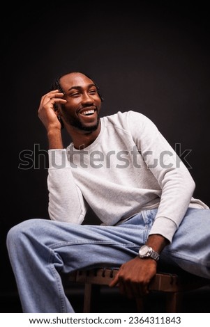 Cheerful and stylish young african american man in sweatshirt and jeans sitting on chair on black background