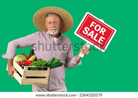 Mature male farmer with wooden box full of different ripe vegetables and FOR SALE sign on green background