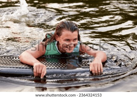Young beautiful sporty girl in a green life jacket swims in the water with a wakeboard in her hands. Happy sportswoman is preparing for a steep wakeboard ride. Active lifestyle, healthy hobby.