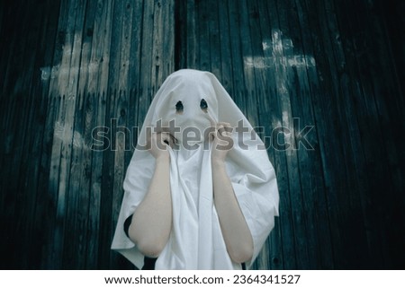 A woman draped in a white ghostly robe with holes for eyes, standing in front of a wooden church adorned with crosses on its doors. The satanism and occultism. The cults and religions. Royalty-Free Stock Photo #2364341527