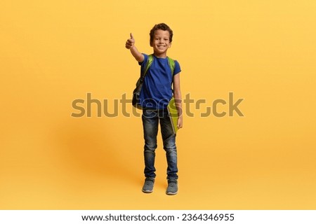 Happy cute preteen black schoolboy with backpack and exercisebook showing thumb up and smiling isolated on yellow studio background. Cheerful african american kid going to school, copy space