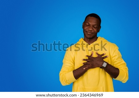 Positive young black guy with closed eyes presses hands to chest, make thanks gesture, isolated on blue background, studio. Gratitude, love sign, human emotions, ad and offer, lifestyle
