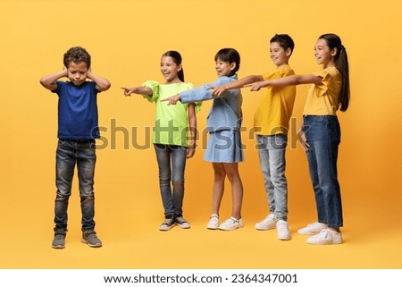 Offended upset preteen boy suffering from bullying from children. Laughing diverse kids pointing at guy covering his ears, isolated on yellow background. Bullying problem among school children Royalty-Free Stock Photo #2364347001