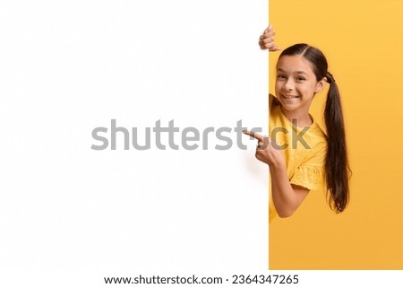 Positive pretty adorable kid girl teenager pointing at huge white empty advertising board isolated on yellow background. Child showing nice advertisement or inspirational slogan text, copy space Royalty-Free Stock Photo #2364347265