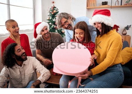 Surprised girl opening Christmas presents at home. Happy and excited family wearing Santa Claus hat gathered together for gift exchange. Positive and joyful domestic relationships in winter vacations.