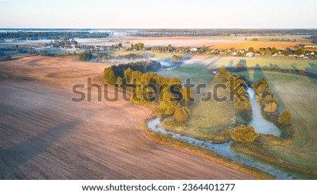 Warm sunny September morning. Autumn beautiful fog landscape. Small river curves in trees. Bright sunny misty riverside. Scenic view of rural sunrise. Village in Agriculture fields. Europe, Belarus