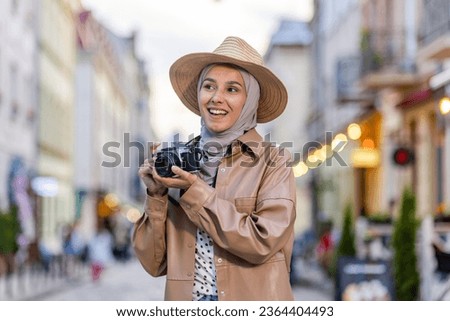 Young beautiful woman walking in the evening city in hijab, tourist with camera and wearing a hat inspects the historical city smiling with satisfaction, Muslim woman on a trip.