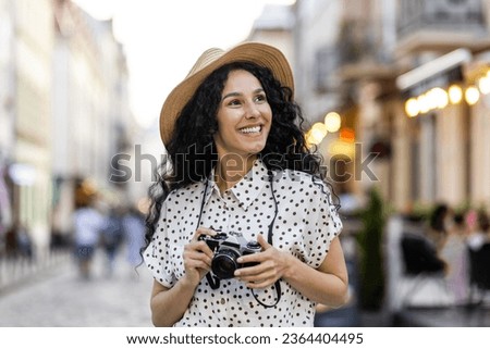 Young beautiful hispanic woman with curly hair walking in the evening city with a camera, female tourist on a trip exploring historical landmarks in the city. Royalty-Free Stock Photo #2364404495