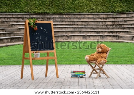 Back to school soon. In the open air there is a wooden board with inscriptions, and a yellow teddy bear sits on a small chair. There are books nearby. Postcard, congratulations, wishes