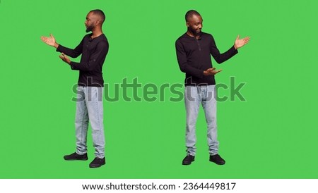 African american man creating advertisement in studio, pointing to left or right sides for ad presentation. Young male model in jeans and shirt presenting something on full body greenscreen.