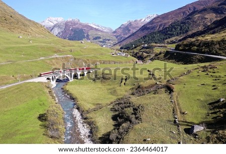A train of Matterhorn Gotthard Bahn travels across a viaduct over a stream in the green valley between alpine mountains on a beautiful sunny day, in Hospental village near Andermatt, Uri, Switzerland Royalty-Free Stock Photo #2364464017