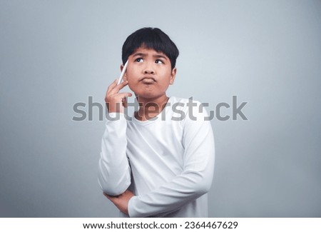 A boy in a white shirt is holding a white pen on a white background. Shows thinking, pondering, and considering options.
