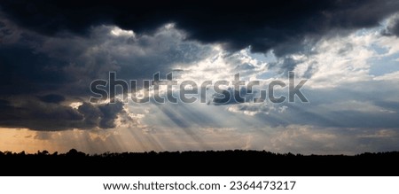 Sunrays coming out of a mix of white and black clouds with a forest silhouette, panorama Royalty-Free Stock Photo #2364473217