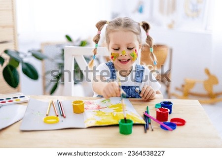 a little child girl paints a drawing in an album and smiles with a dirty face and hands, a happy child draws a picture at home at the table.