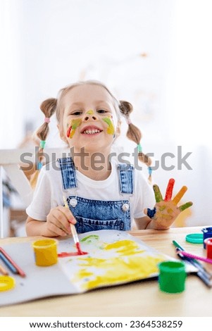 a little child girl paints a drawing in an album and smiles with a dirty face and hands, a happy child draws a picture at home at the table.