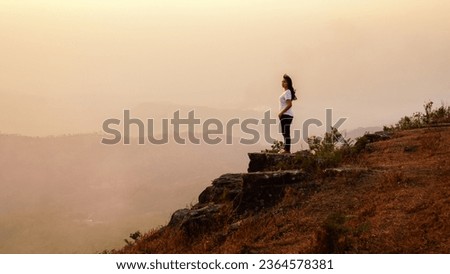 Woman hiker standing on the edge of cliff at Phu Bak Dai, Loei, Thailand.. Female reaching summit enjoying amazing sunset in the mountains. Backlight sunlight with beautiful lens flare and sunbeam.