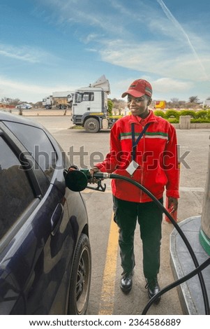 african american attendant in uniform at the gas station, filling with petrol a small car, nozzle is in the tank Royalty-Free Stock Photo #2364586889
