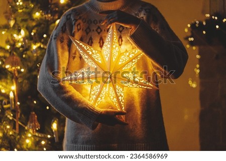 Hands in cozy sweater holding golden illuminated christmas star on background of stylish decorated christmas tree with golden lights in evening room. Atmospheric magical eve. Merry Christmas!