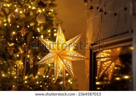 Atmospheric christmas eve. Stylish christmas illuminated star, decorated christmas tree with golden lights and festive decor on fireplace mantel in scandinavian room. Merry Christmas! Royalty-Free Stock Photo #2364587013