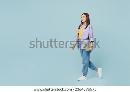 Full body side view young happy IT woman wear purple shirt yellow t-shirt casual clothes hold closed laptop pc computer isolated on plain pastel light blue background studio portrait Lifestyle concept
