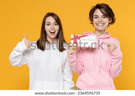 Excited two young women friends 20s wearing casual white pink hoodies standing pointing index finger on gift certificate showing thumb up isolated on bright yellow color background studio portrait