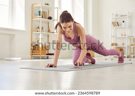Portrait of a young sporty athletic girl or fitness trainer woman wearing sportswear exercising on the floor on yoga mat at home having online training by mobile phone. Fitness and workout concept. Royalty-Free Stock Photo #2364598789