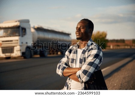 Serious look. With arms crossed. Black man is standing on the road with truck on it.