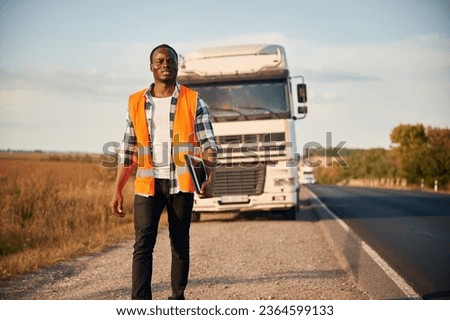 Front view. Black man is standing on the road with truck on it.