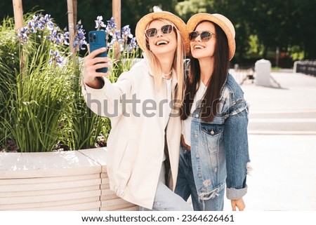 Two young beautiful smiling hipster female in trendy summer clothes. Carefree women posing in the street. Positive models having fun at sunny day. Cheerful and happy. In hat, sunglasses. Take selfie