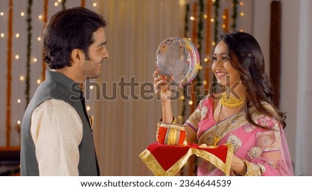 Young Indian couple happily performing Karwa Chauth traditions in traditional wear. Beautiful wife looking at the moon through a sieve and then to her good looking husband, following Karwa Chauth r... Royalty-Free Stock Photo #2364644539