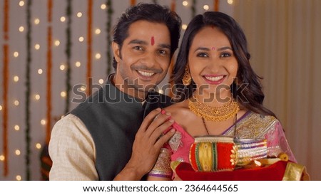 Indian husband-wife posing and smiling while looking into the camera - celebrating Karwa Chauth festival. Newly married couple celebrating their first Karwa Chauth festival - Dressed in traditional... Royalty-Free Stock Photo #2364644565