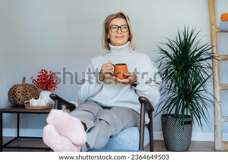 Middle aged woman relaxing with cup of hot drink in scandy style hygge interior home with fall mood interior decor. Lady dreaming, enjoy calm mood without stress, wellbeing alone. Cozy autumn at home. Royalty-Free Stock Photo #2364649503