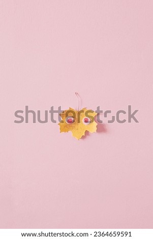 Creative yellow fall leaf with pink eyes on a pink pastel background. Minimal holiday, autumn, thanksgiving, or Halloween concept.