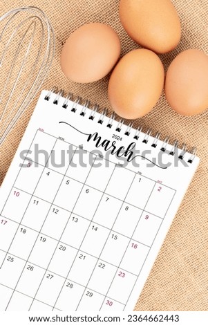 March 2024 Desk calendar and fresh eggs with egg whisk on burlap background.