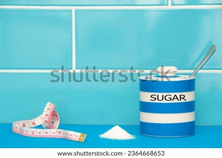 Sugar concept. A tape measure sits next to a pile of white sugar and sugar pot. A blue themed picture with good copy space. Diet and lifestyle picture. Selective focus on the front of the cup. 